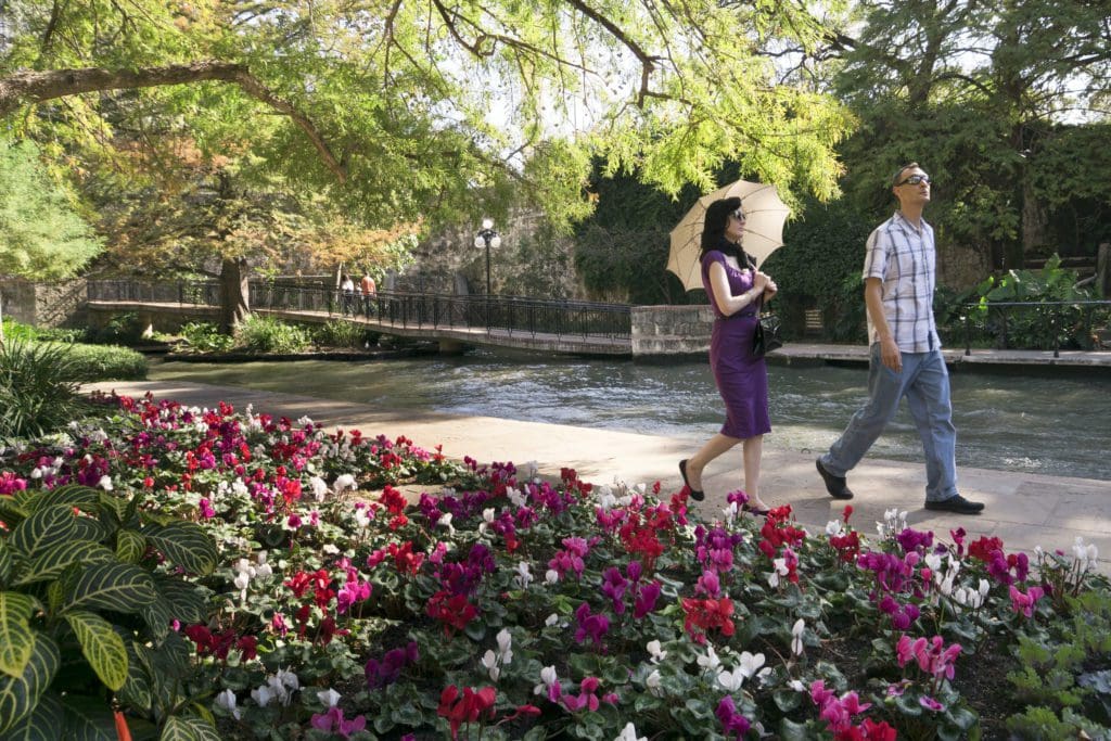 A man and a woman stroll along the River Walk bank with colorful flowers in the foreground
